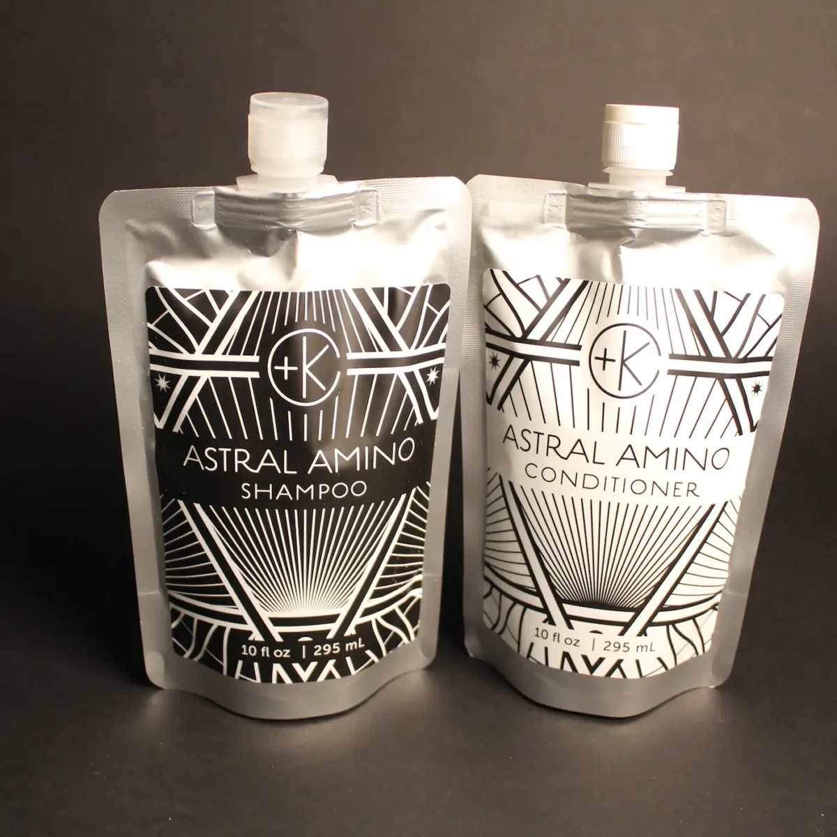 2 silver packages of infinitely recyclable aluyminum packaging show the new astral amino and conditioner from c+k. the shampoo label is black and the conditioner label is white. they have white flip top lids and a wider base to stnad on narrowing toward the top.
