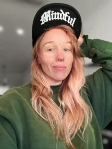 Erin wears an olive green carhartt crewneck and a black flip bil hat that says mindful in an old english homage.