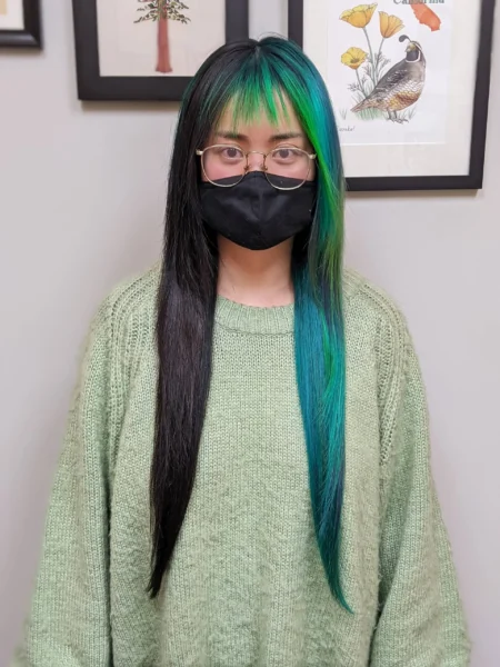 Person with long straight black and teal hair with matching teal bangs, wearing glasses, a black face mask, and a loose green sweater, standing in front of framed botanical art.
