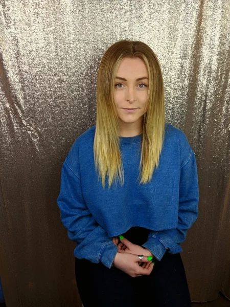 Woman with straightened blonde hair wearing a blue sweater, seated against a sparkly sequin backdrop.