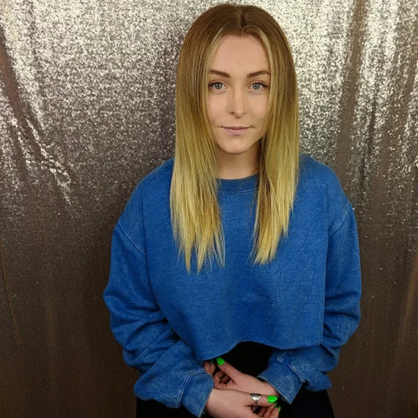 Woman with straightened blonde hair wearing a blue sweater, seated against a sparkly sequin backdrop.