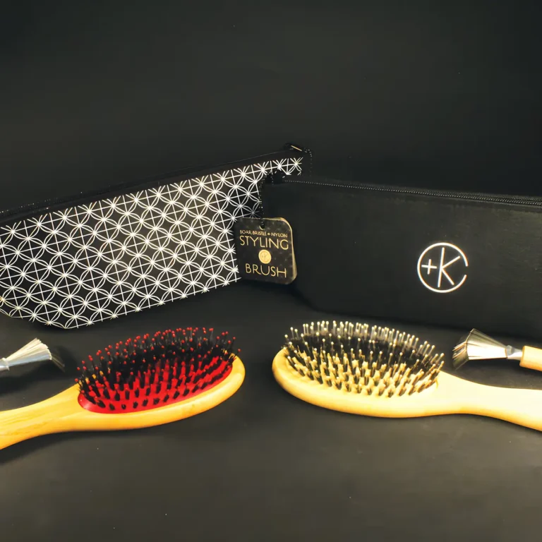 a red brush and a yellow brush with matching bamboo handles and boar bristles sit in front of their black felt carrying cases and cleaning rakes on a black background.