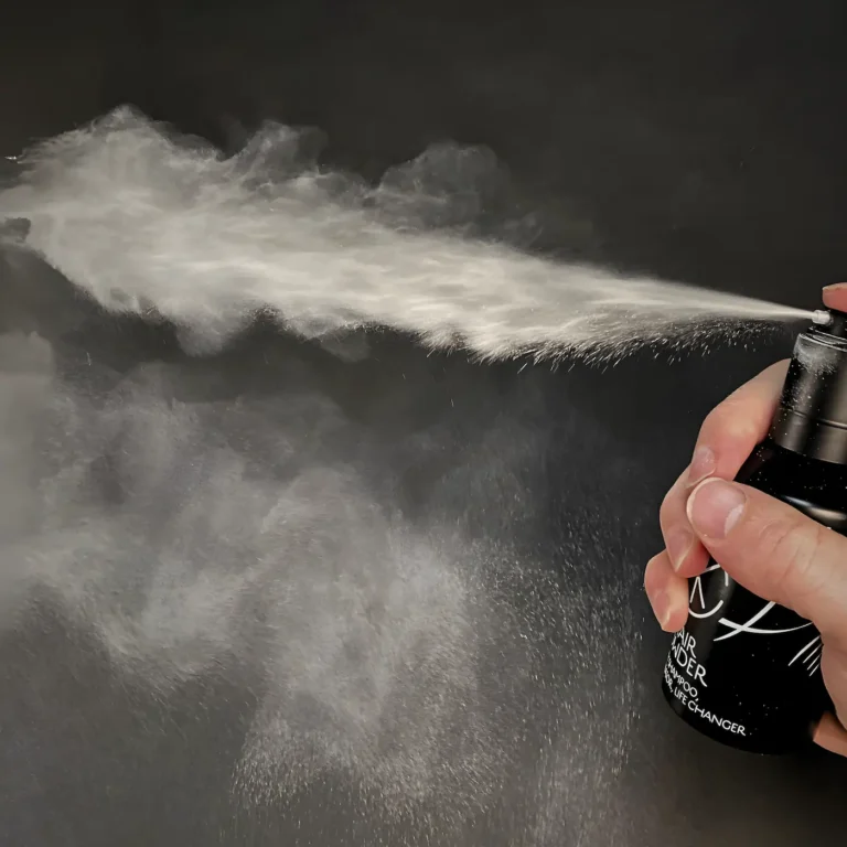 a hand sprays a bottle of powder that has a hella long puff and a fine powder spray. the hand is white and the background is black amd the bottle is black.