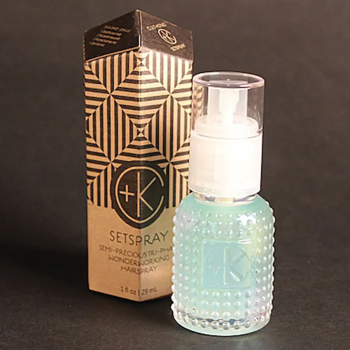 a mini glass bottle with a white spray lid and a malachite colored fluid inside stands in front of an obelisk cardboard package.