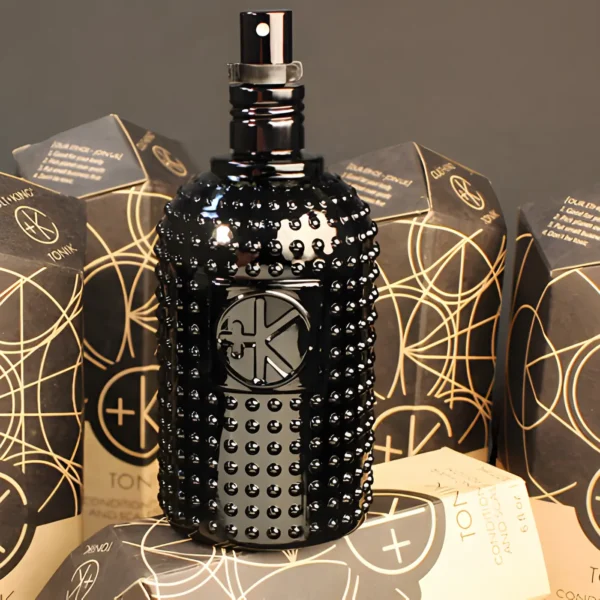 a black glass bottle with glass dots and a black sprayer with a lid on it sits on a black background in front of it's obelisk cardboard packaging.