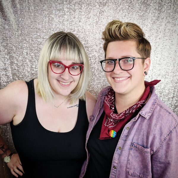 Two people smiling in front of a sparkly backdrop, one with a blonde bob and bangs wearing red glasses, and the other with short styled brown hair, black glasses, and a red bandana with a rainbow pin.