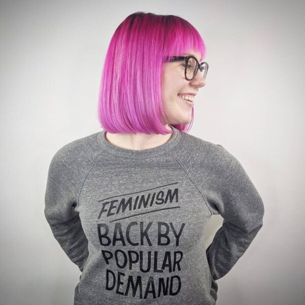 Person with a bright magenta bob haircut wearing glasses and a gray sweatshirt that reads “Feminism Back by Popular Demand,” smiling and looking to the side.