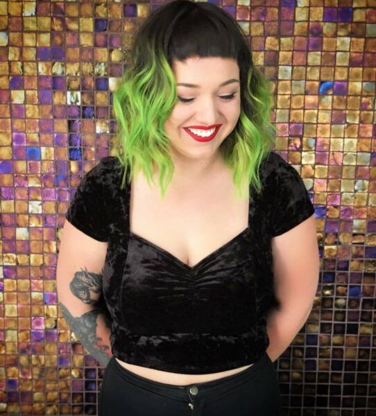 Person with bright green wavy bob haircut, short bangs, and a black velvet crop top, smiling against a colorful mosaic tile background.