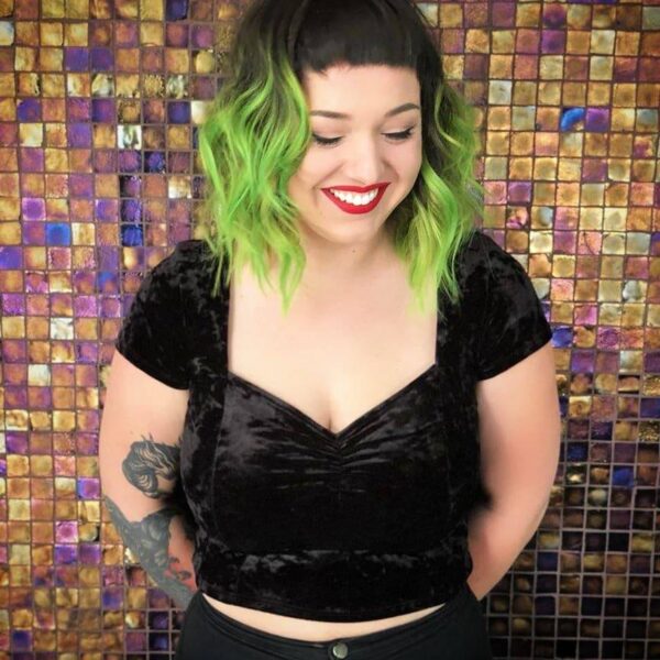 Person with bright green wavy bob haircut, short bangs, and a black velvet crop top, smiling against a colorful mosaic tile background.