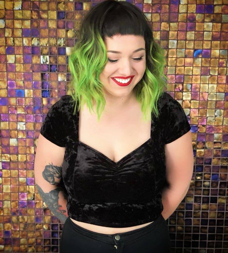 Person with bright green wavy bob haircut, short bangs, and a black velvet crop top, smiling against a colorful mosaic tile background.