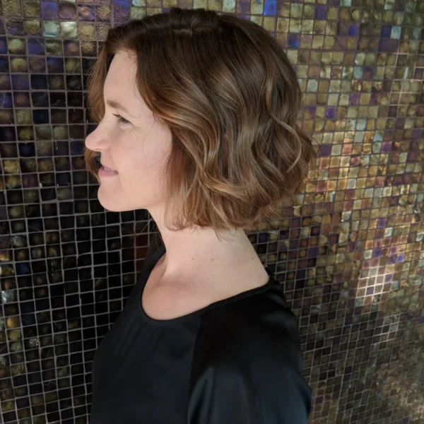 a woman stands shwing her profile in front of a mosaic wall, her hair is rich brunette in a french bob, styled in a gentle wave.