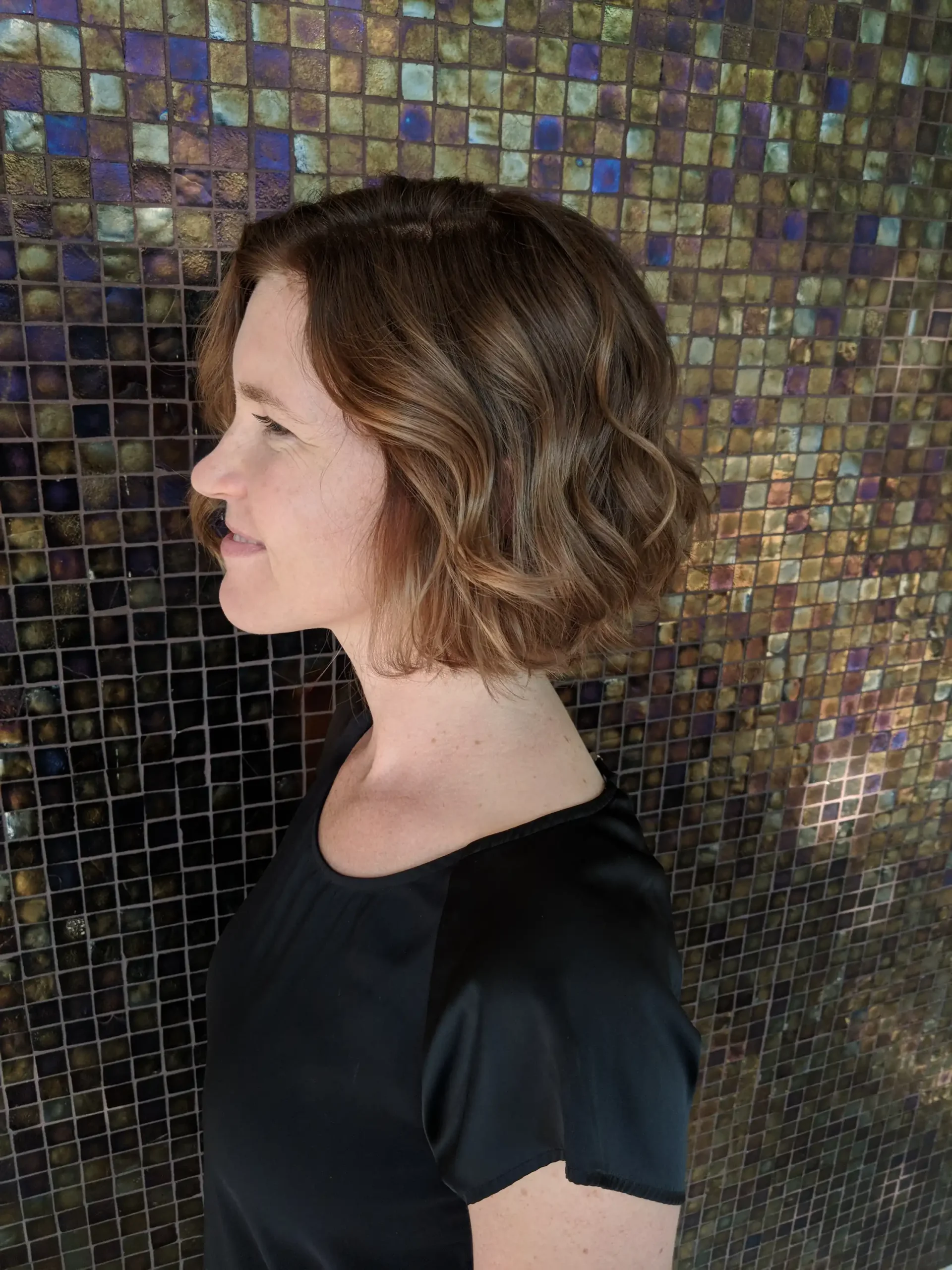 a woman stands shwing her profile in front of a mosaic wall, her hair is rich brunette in a french bob, styled in a gentle wave.