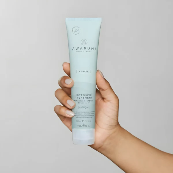 a hand holding a white bottle of conditioner on a plain light grey background. it's a reparative treatment for damaged hair