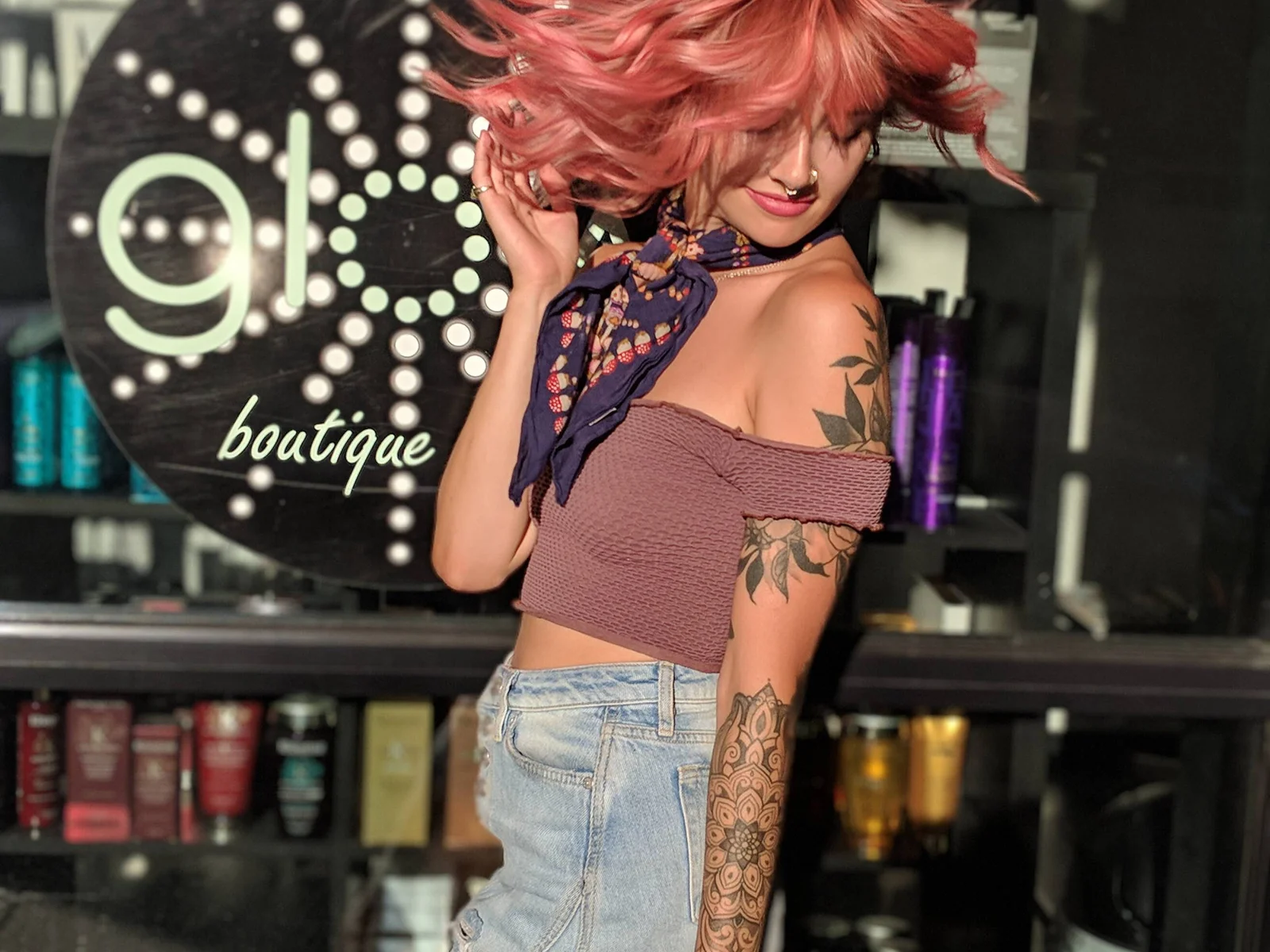 Person with wavy pink hair in motion, wearing a crop top, scarf, and denim shorts, posing in front of a "glo boutique" sign with tattooed arm visible.