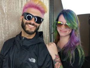 a man with a short haircut dyed pink at the roots and yellow at the tips wears sunglasses and stands next to a woman with oil slickish long hair and she also wears sunglasses. there is a champagne colored backdrop behind them.