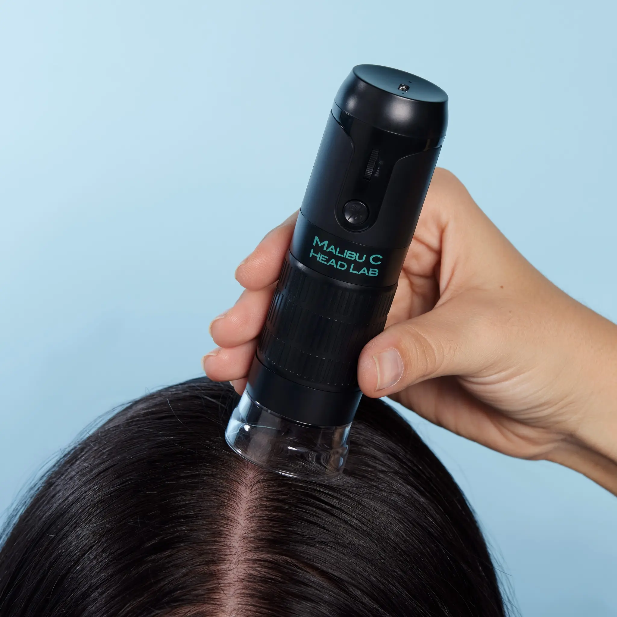 a hand holding a scalp camera to a persons scalp. the hair is dark and the background is a light blue.