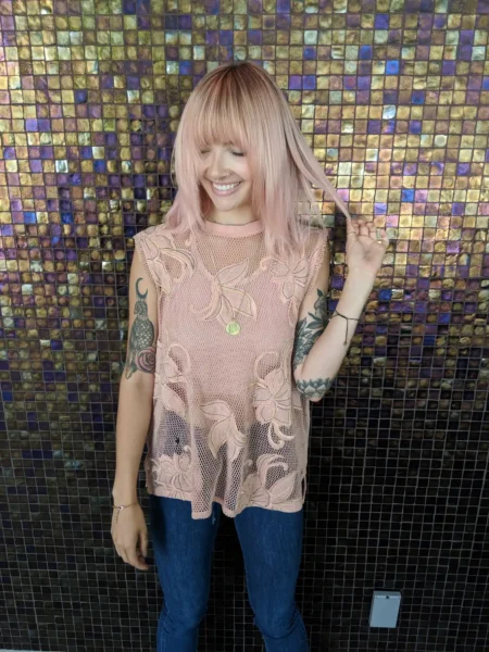 a woman with soft pink blonde hair stands in front of a mosaic wall after receiving a hair repair treatment called the seasonal reset. she is smiling and looking down and touching her hair which is very soft and shiny