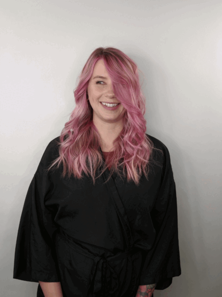 a woman with dimensional pink hair stands in front of a white wall and wears a black robe. she is smiling and her hair is curled.
