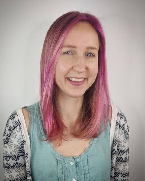 Smiling person with soft pink straight hair, wearing a teal top with a patterned cardigan, posing against a plain background.
