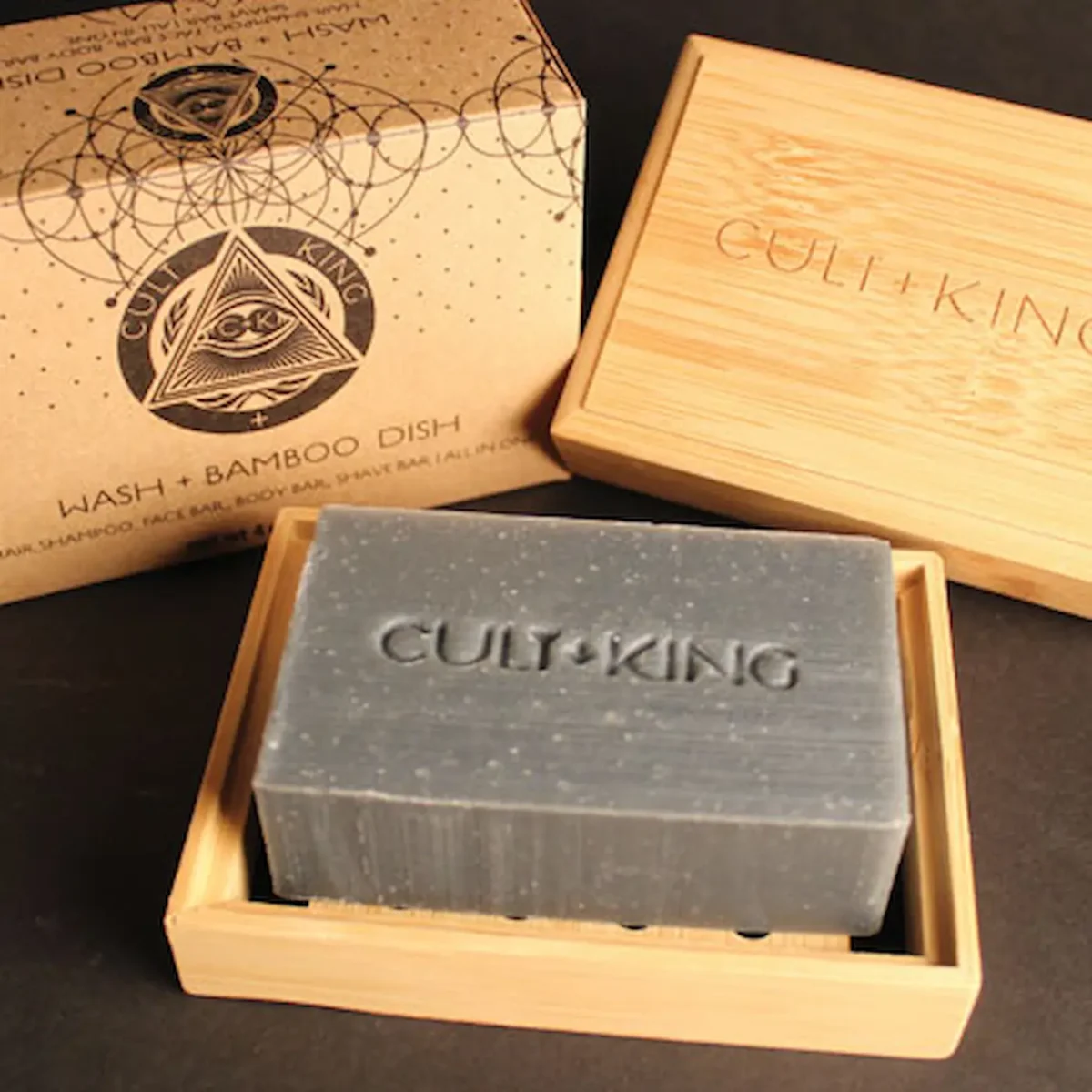 a charcoal grey bar of soap sits in a light colored bamboo soap dish and the packaging box with alchemical symbols on cardboard sits behind it. Cult + King is embossed in the bar.