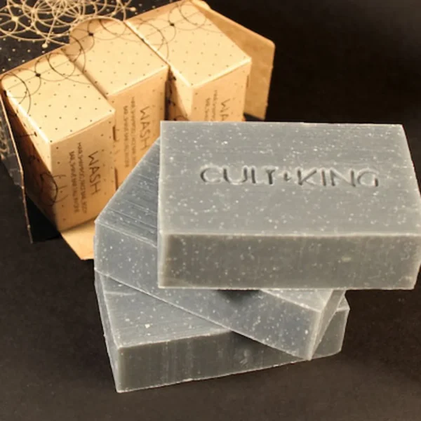 3 charcoal grey bars of shampoo are stacked diagonally with Cult + King stamped in to the bars with the light brown cardboard boxes behind them and they are on a black background.