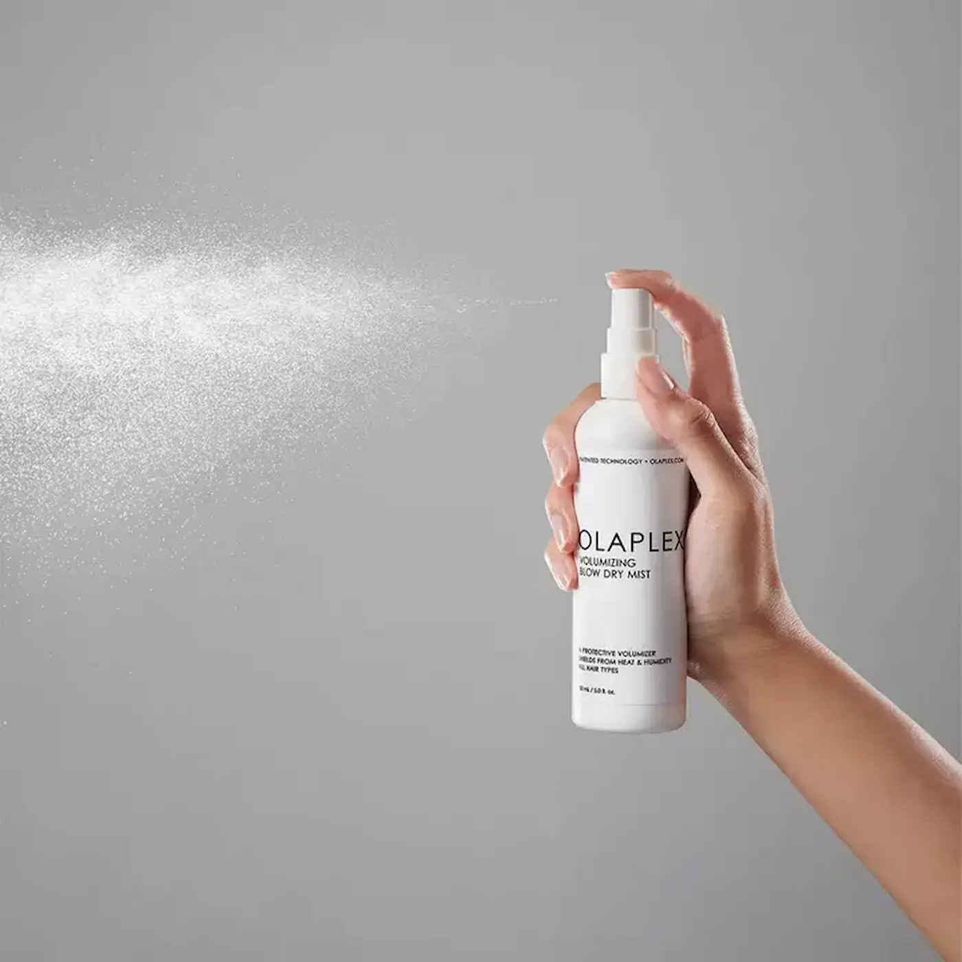 Hand spraying Olaplex Volumizing Blow Dry Mist against gray background.