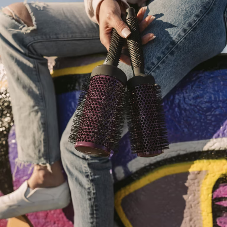 a hand holds 2 different sized round brushes from ergo. the super gentle round brushes have a purple colorway on the barrel and black bristles and handle.