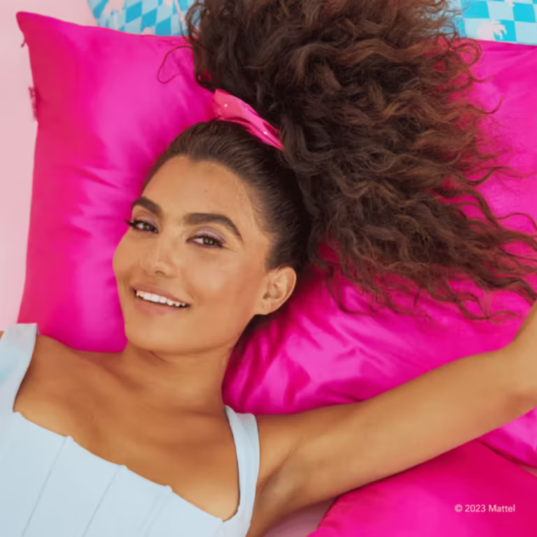 a beautiful tan woman with dark brown frizzy hair in a hot pink scrunchie lays on a luxurious hot pink satin pillowcase in a tank top
