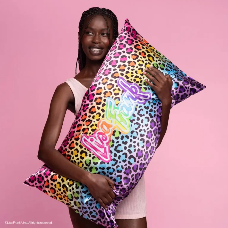 a woman stands in front of a pnk backdrop ans she holds her king sized pillow with a rainbow leopard lisa frank logo