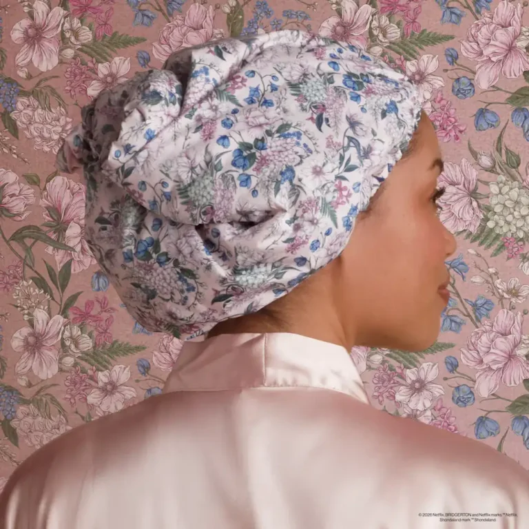 a woman wears the wisteria satin lined shower cap in front of classic bridgerton floral wallpaper