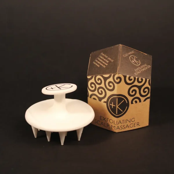 a black background shows a white silicone scalp brush next to it's obelisk cardboard packaging