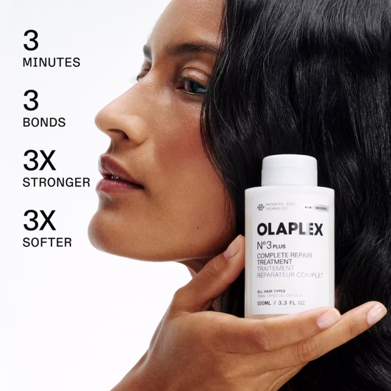 a woman holds a white bottle of olaplex no 3 plus in her hand and looks away demurely